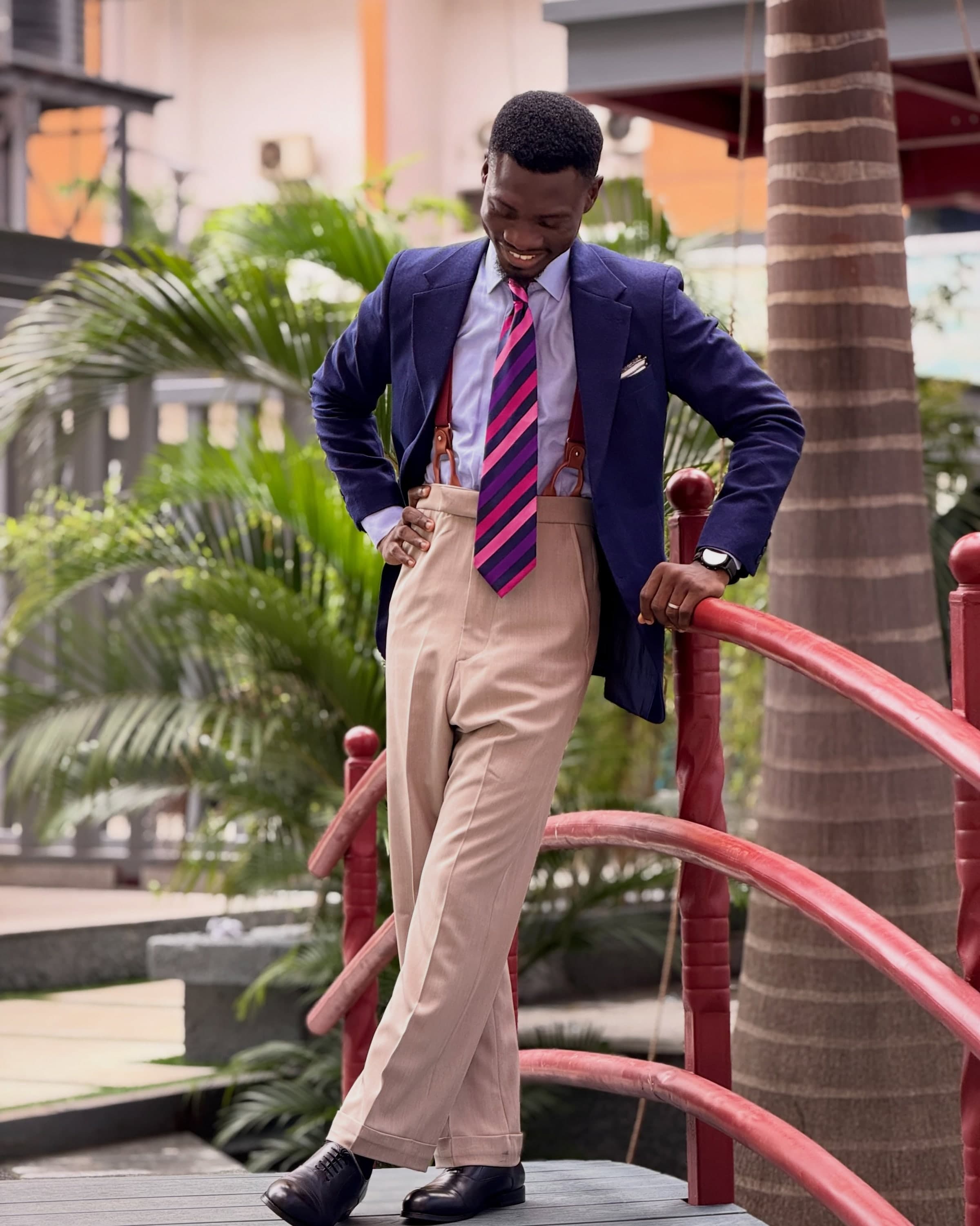 A man in a navy blazer, striped tie and braces leaning on a red bridge railing