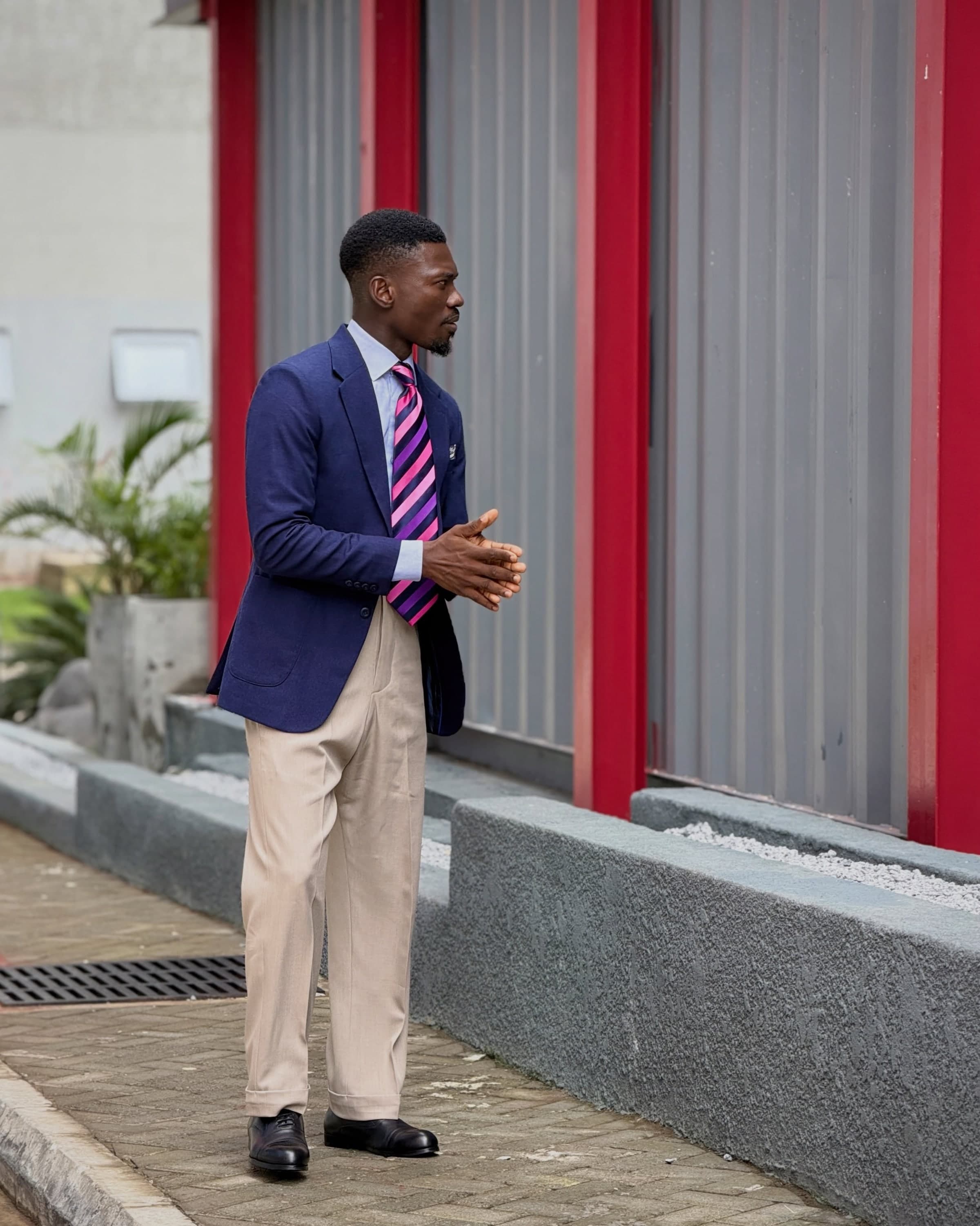 A man in a navy blazer and cream trousers standing in profile beside red columns
