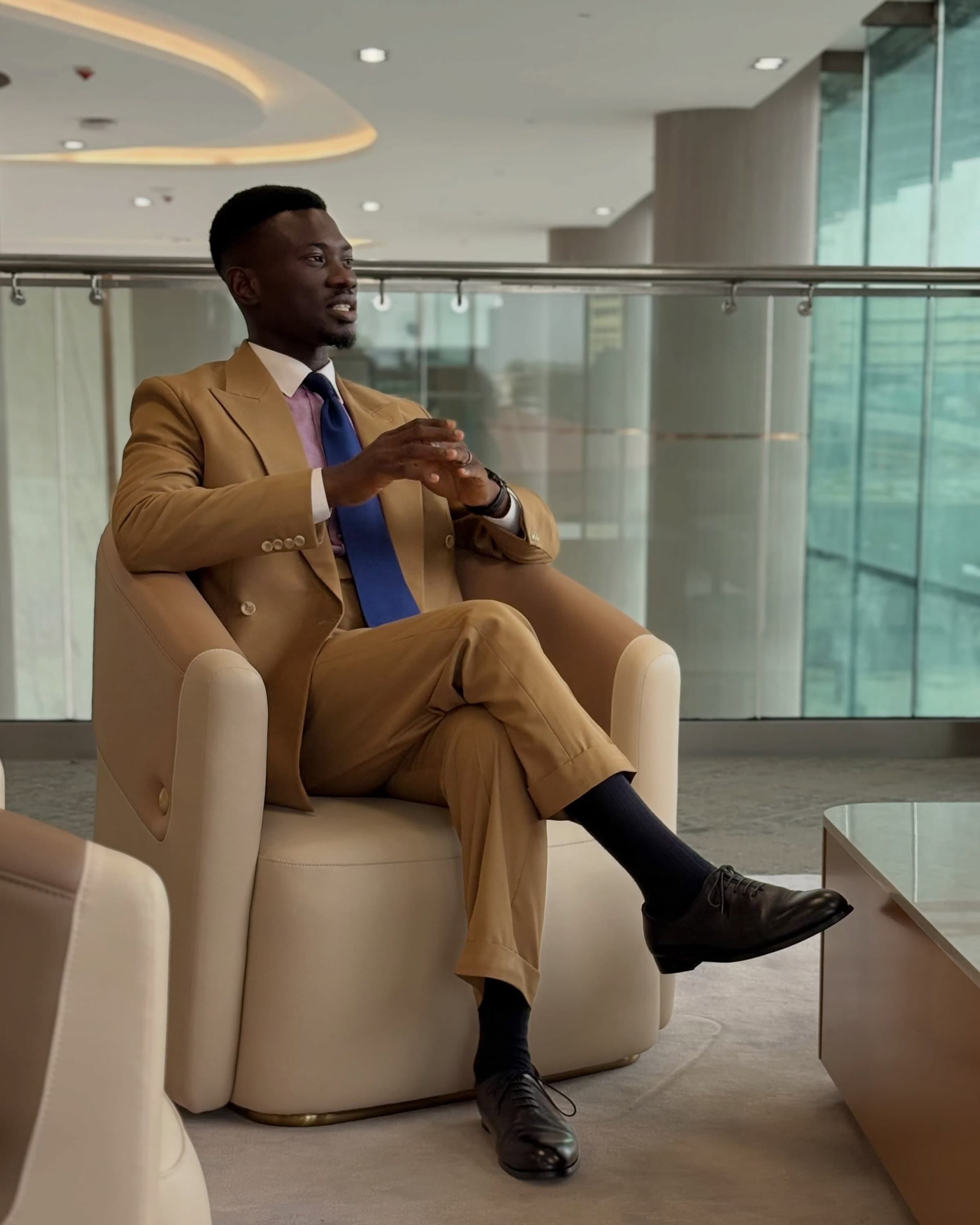 A man in a tan double-breasted suit seated in profile in a bright atrium