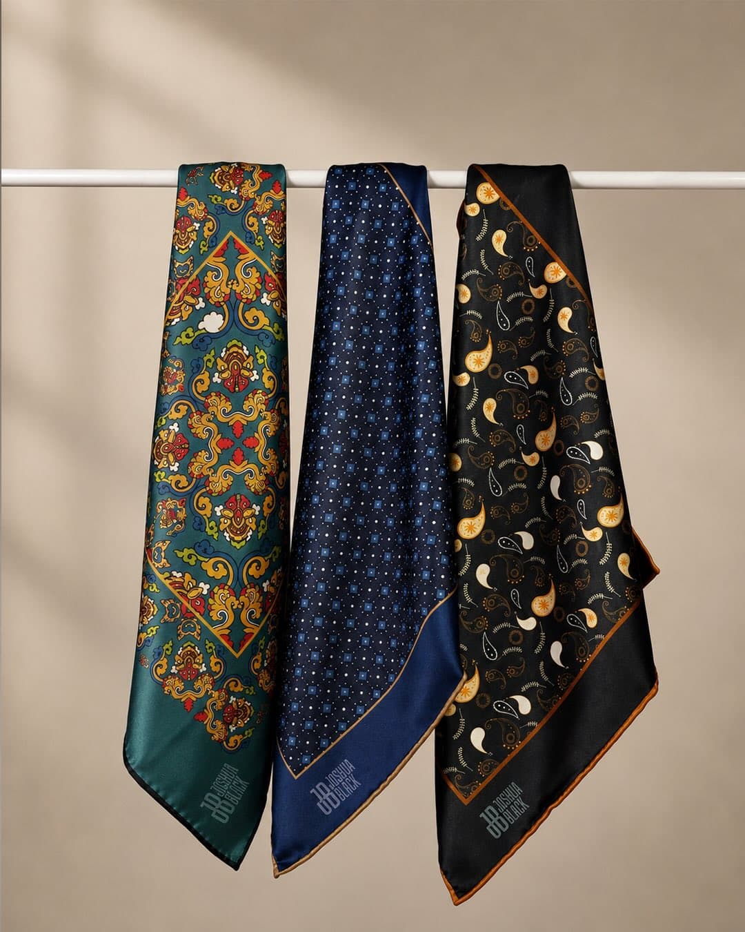 Three pocket squares hung over a rail: emerald medallion, navy diamond and black paisley